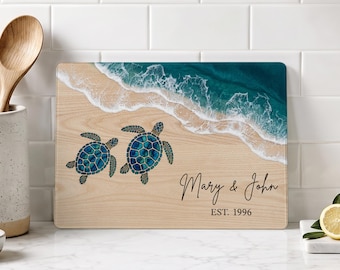 May include: A rectangular wooden cutting board featuring a printed design of two blue sea turtles on a sandy beach with a teal ocean wave. The text "Mary & John" and "EST. 1996" is printed in black script and serif font on the lower right side.