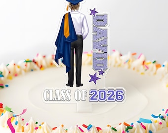 May include: A graduation cake topper featuring a figure in a cap and gown, with the name "DAVID" and "CLASS OF 2026" in purple. The cake is decorated with white frosting, sprinkles, and colorful ribbons.