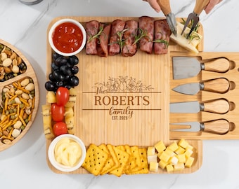May include: A bamboo charcuterie board with the engraved text "The ROBERTS family EST. 2025". The board is filled with snacks, including crackers, cheese, grapes, and tomatoes. Cheese knives and forks are included.