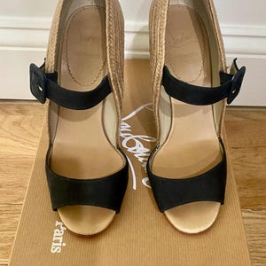 May include: A pair of black and tan espadrille wedge sandals with a buckle closure. The sandals are on a brown box with the brand name "Christian Louboutin Paris" printed on the side.