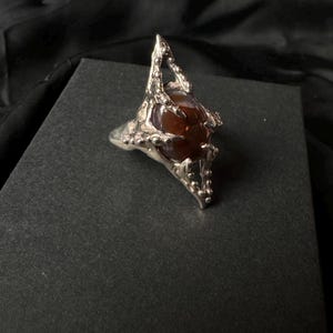 Könnte beinhalten: Ein silberner Ring mit einem braunen Edelstein, der in einem klauenartigen Design gefasst ist.