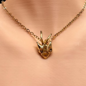 Puede incluir: Un collar dorado con un colgante con forma de cabeza de insecto. El colgante tiene detalles intrincados y cuelga de una fina cadena. El collar se muestra sobre un fondo neutro, resaltando la joya.