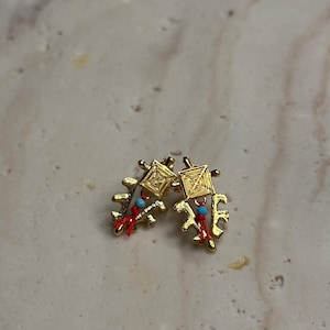 May include: A pair of gold-colored stud earrings. Each earring features a square top with a woven design, a red tassel, and a small turquoise bead. The earrings have a unique, abstract shape.