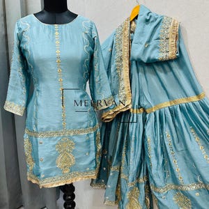 Może przedstawiać: Jasnoniebieski, haftowany zestaw tuniki i pasujących spodni z rozszerzanymi nogawkami. Tunika posiada złote hafty wzdłuż dekoltu, rękawów i dołu. Spodnie mają rozszerzany krój z podobnymi złotymi haftami. Zestaw jest prezentowany na manekinie i wieszaku.