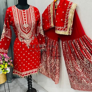 Puede incluir: Un vibrante conjunto bordado en rojo que incluye una túnica, pantalones palazzo y una bufanda a juego. La túnica está adornada con intrincados bordados dorados, y los pantalones palazzo tienen un diseño amplio y fluido. El conjunto se exhibe en un maniquí.