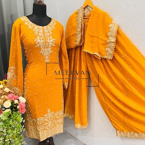 May include: Mustard yellow embroidered salwar kameez set. The long-sleeved tunic and matching pants are adorned with intricate gold embroidery and small pearl accents. A matching shawl with gold trim completes the ensemble. The text "MEERVAN ENTERPRISE" is visible.