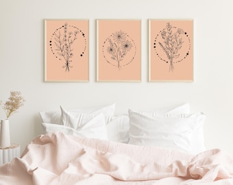 Op de afbeelding: Drie ingelijste botanische kunstprints. Elke print toont een zwarte lijntekening van een bloemstuk op een perzikachtergrond, omcirkeld door een stippellijn. De frames zijn van licht hout. De prints worden weergegeven boven een bed met witte kussens en een roze deken.