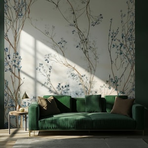 Puede incluir: Un sofá de terciopelo verde con patas doradas y varios cojines. El fondo presenta un mural de ramas de árboles con hojas azules. A la izquierda hay una pequeña mesa auxiliar con una lámpara y objetos decorativos.
