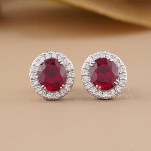 May include: A pair of oval-cut deep red ruby stud earrings, each surrounded by a halo of small, clear, round-cut white diamonds set in a polished silver-toned metal frame. The earrings feature a classic prong setting for each stone.