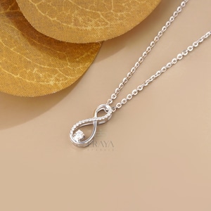 Peut inclure: Un collier argenté avec un pendentif en forme de symbole infini. Le pendentif est orné de petites pierres claires sur un côté et d'une pierre claire plus grande dans la boucle inférieure. Le texte ERAYA SPARKLE est visible.