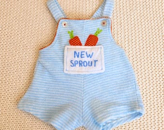 May include: Light blue baby overalls with horizontal stripes. The overalls have a pocket with the words "NEW SPROUT" embroidered on it. Two carrot appliqués are above the pocket. The overalls have a red trim.