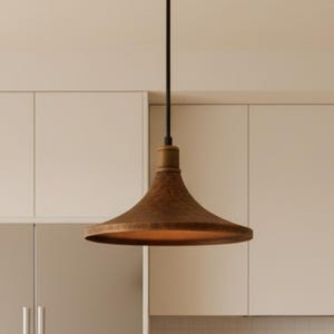 May include: A bronze-coloured, bell-shaped pendant light hangs from a black cord in a kitchen. The light fixture has a rustic style. The kitchen features white cabinets and a stainless steel fridge.