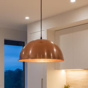 May include: A copper-colored pendant light fixture with a dome-shaped shade hangs from the ceiling in a modern kitchen. The light illuminates the kitchen island and surrounding area. The kitchen features white cabinets and a light-colored countertop.