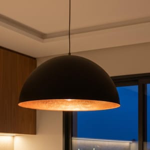 May include: A black pendant light with a copper-colored interior hangs from the ceiling. The light fixture is dome-shaped and suspended above a kitchen countertop. The kitchen features wooden cabinets and a window with a view of the night sky.