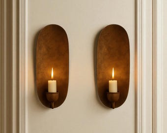 Op de afbeelding: Twee wandkandelaars met een warme, metalen afwerking. Elke kandelaar bevat een brandende, ivoorkleurige kaars. De kandelaars zijn bevestigd aan een neutraal gekleurde muur, wat een gezellige en uitnodigende sfeer creëert. Het ontwerp is eenvoudig maar elegant.