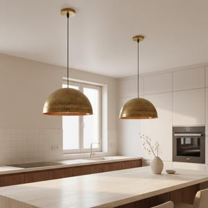 Puede incluir: Dos l&aacute;mparas colgantes de lat&oacute;n con forma de c&uacute;pula cuelgan del techo. Las luces tienen una superficie texturizada y un tono dorado c&aacute;lido. Est&aacute;n suspendidas sobre una isla de cocina con una encimera de color claro.
