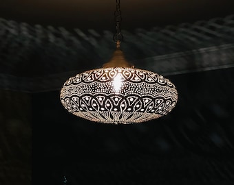 May include: An oval-shaped, gold-colored pendant light fixture with intricate cut-out patterns. The light is suspended from a chain and illuminates a dark room, casting patterned shadows on the ceiling and walls. The fixture has a warm, inviting glow.