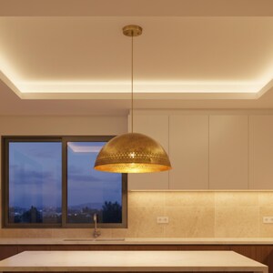 Puede incluir: Una l&aacute;mpara colgante dorada en forma de c&uacute;pula que cuelga del techo. La luz tiene una banda decorativa perforada alrededor del borde inferior. El entorno de la cocina incluye una ventana, armarios y una encimera.