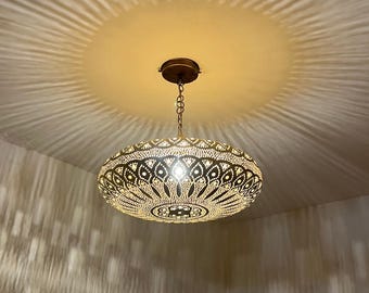 May include: Ornate, gold-toned pendant light with intricate cut-out patterns. The light fixture is suspended from the ceiling by a chain and casts a decorative pattern of light and shadow on the ceiling and walls.