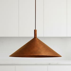 Puede incluir: Una lámpara colgante de color cobre con una pantalla cónica. La luz cuelga de un cable oscuro y tiene una parte superior cilíndrica. La lámpara es de metal y tiene un aspecto cálido y rústico. El fondo es una pared blanca.