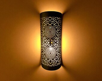 May include: A cylindrical wall sconce with a black, patterned metal shade. The shade features intricate cut-out designs and two circular patterns. The sconce emits a warm, inviting light, casting shadows on the surrounding wall.