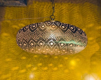 May include: A decorative, oval-shaped pendant light fixture. The metal shade is a bronze color and features a pattern of small dots and diamond shapes. The light is suspended from a chain and casts a warm glow on the yellow wall.