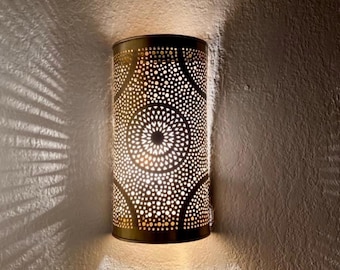 May include: A cylindrical wall sconce with a dark bronze finish. The sconce features a pattern of small, evenly spaced holes that emit light, creating a decorative shadow effect on the wall. The design includes circular and geometric shapes.