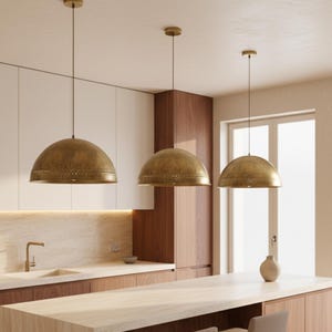 Puede incluir: Tres l&aacute;mparas colgantes de lat&oacute;n con forma de c&uacute;pula cuelgan sobre una isla de cocina. Las luces tienen un acabado met&aacute;lico c&aacute;lido y est&aacute;n suspendidas del techo. La cocina presenta gabinetes de madera clara y una paleta de colores neutros.