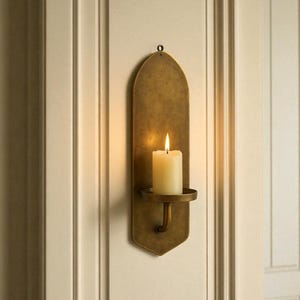 May include: A wall-mounted candle holder with a lit, ivory-colored candle. The holder is made of aged brass and has a decorative, elongated shape. The candle holder is mounted on a cream-colored wall.
