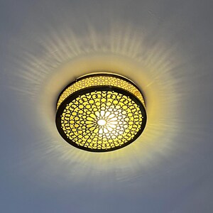 Pode incluir: Uma luminária de teto circular com um design ornamental vazado. A luminária tem uma estrutura de metal escuro e emite uma luz amarela quente, criando sombras. O design lembra a arte marroquina ou islâmica.