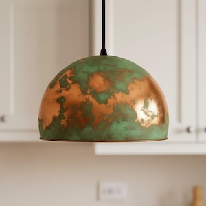 Puede incluir: Lámpara colgante en forma de cúpula con acabado de pátina de cobre y verde. La lámpara cuelga de un cable negro sobre un fondo blanco. El color cobre es visible en parches.