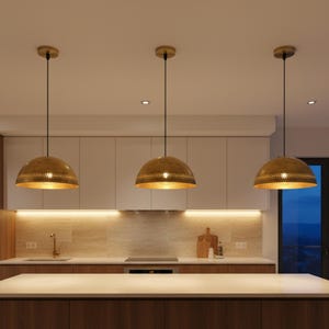 Puede incluir: Tres l&aacute;mparas colgantes doradas con un dise&ntilde;o perforado cuelgan sobre una isla de cocina. Las l&aacute;mparas tienen forma de c&uacute;pula y est&aacute;n suspendidas por cables negros. La cocina cuenta con armarios blancos y una encimera clara.