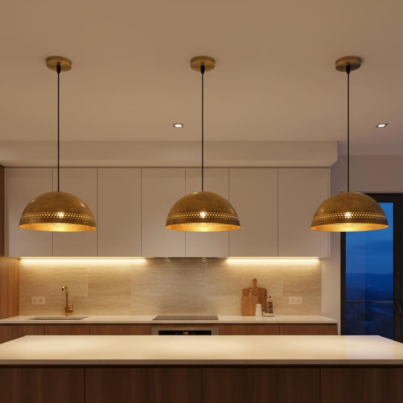 Puede incluir: Tres l&aacute;mparas colgantes doradas con un dise&ntilde;o perforado cuelgan sobre una isla de cocina. Las l&aacute;mparas tienen forma de c&uacute;pula y est&aacute;n suspendidas por cables negros. La cocina cuenta con armarios blancos y una encimera clara.