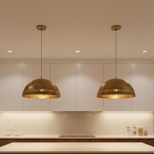 Puede incluir: Dos l&aacute;mparas colgantes de lat&oacute;n con forma de c&uacute;pula cuelgan del techo. Las l&aacute;mparas tienen un dise&ntilde;o perforado en el borde inferior y un interior dorado. Las luces est&aacute;n suspendidas sobre una isla de cocina.