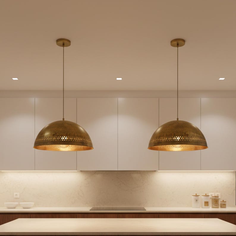 Puede incluir: Dos l&aacute;mparas colgantes de lat&oacute;n con forma de c&uacute;pula cuelgan del techo. Las l&aacute;mparas tienen un dise&ntilde;o perforado en el borde inferior y un interior dorado. Las luces est&aacute;n suspendidas sobre una isla de cocina.