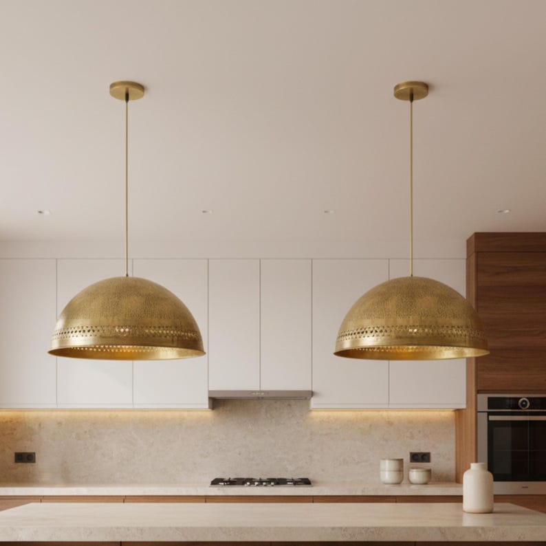 Puede incluir: Dos l&aacute;mparas colgantes doradas con forma de c&uacute;pula cuelgan del techo. Las l&aacute;mparas tienen una superficie texturizada y una fila de peque&ntilde;os agujeros cerca de la parte inferior. La cocina cuenta con gabinetes blancos y una encimera de color claro.