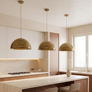 Puede incluir: Tres l&aacute;mparas colgantes de lat&oacute;n con forma de c&uacute;pula cuelgan en una cocina. Las l&aacute;mparas tienen una superficie texturizada y est&aacute;n suspendidas del techo. La cocina cuenta con armarios blancos y una encimera clara.