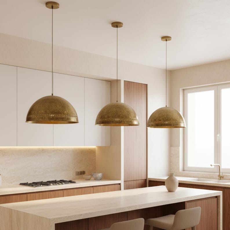 Puede incluir: Tres l&aacute;mparas colgantes de lat&oacute;n con forma de c&uacute;pula cuelgan en una cocina. Las l&aacute;mparas tienen una superficie texturizada y est&aacute;n suspendidas del techo. La cocina cuenta con armarios blancos y una encimera clara.