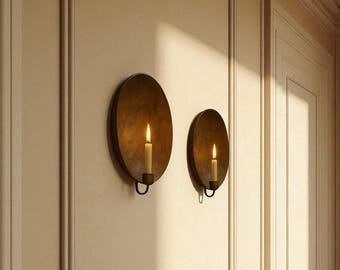 May include: Two antique-style wall sconces with lit candles. The sconces are circular, made of a dark metal, and mounted on a light-colored wall. The candles have a warm, flickering glow, creating a cozy ambiance. The sconces are a decorative lighting fixture.