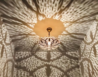 May include: Ornate, gold-colored pendant light with intricate cut-out patterns, casting a decorative light and shadow display on the ceiling and walls. The light fixture is suspended from a brass-toned canopy.