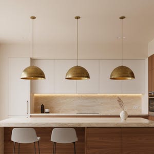 Puede incluir: Tres l&aacute;mparas colgantes de lat&oacute;n sobre una isla de cocina. Las l&aacute;mparas tienen forma de c&uacute;pula con superficie texturizada. La cocina cuenta con gabinetes blancos, encimera de m&aacute;rmol y base de isla de madera. Un jarr&oacute;n con una planta est&aacute; en la isla.