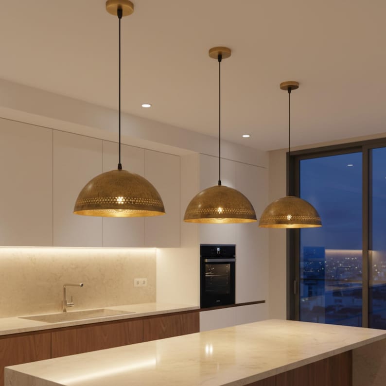Puede incluir: Tres l&aacute;mparas colgantes de lat&oacute;n con un dise&ntilde;o perforado cuelgan sobre una isla de cocina. Las luces tienen forma de c&uacute;pula y est&aacute;n suspendidas por cables negros. La cocina cuenta con gabinetes blancos, una encimera de m&aacute;rmol y un horno empotrado.