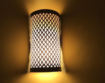 May include: A decorative wall sconce with a cylindrical metal shade featuring a lattice pattern. The sconce emits a warm, inviting light, casting shadows on the surrounding wall. The fixture is mounted on a neutral-colored wall.