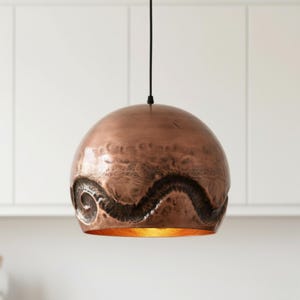 May include: A copper-colored pendant light fixture with a textured surface and a dark, wave-like design. The interior of the light fixture has a golden hue. The light hangs from a black cord, suitable for kitchen or dining room lighting.