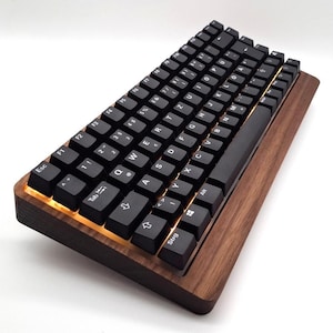 Könnte beinhalten: Eine schwarze mechanische Tastatur mit schwarzen Tastenkappen und weißer Beschriftung liegt auf einer Holzbasis. Die Tastatur hat ein kompaktes Layout und ist nach oben abgewinkelt. Die Holzbasis hat einen warmen, braunen Farbton, und die Tasten sind hintergrundbeleuchtet.