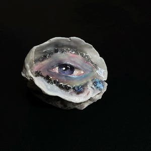 May include: A natural oyster shell featuring a hand-painted eye with a purple iris and detailed lashes. The surrounding area is decorated with iridescent pigments and small, dark, reflective mineral-like accents embedded in a glossy resin finish.