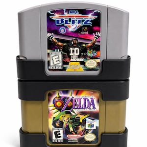 May include: A grey NFL Blitz video game cartridge stacked on top of a gold The Legend of Zelda: Majora's Mask video game cartridge, both held within a black plastic storage stand. Each cartridge features its respective game title and artwork.