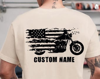Puede incluir: Camiseta color crema con un diseño desgastado de la bandera estadounidense y una silueta de motocicleta. El texto "CUSTOM NAME" está impreso debajo. La camiseta se usa en un garaje, con motocicletas en el fondo.