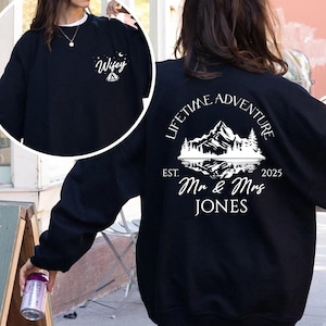 May include: Black sweatshirt with white graphic on the back that reads "Lifetime Adventure" above a mountain scene, "Est. 2025", and "Mr & Mrs JONES". A smaller graphic on the front says "Wifey".