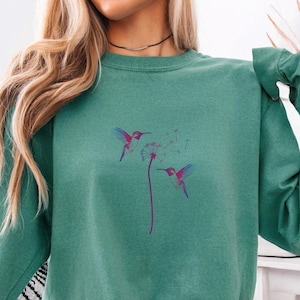 Pu&ograve; includere: Una felpa verde acqua con un disegno di colibr&igrave; e dente di leone. La grafica mostra due colibr&igrave; con ali viola e blu e un becco rosa, che si librano vicino a un dente di leone. La felpa ha maniche lunghe e girocollo.